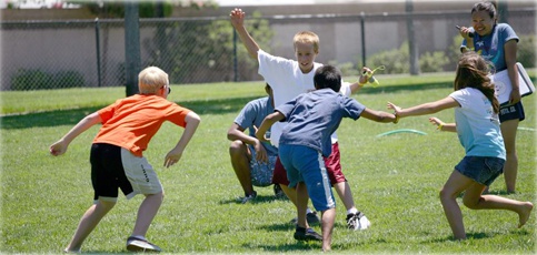 Photo shows a group of four children and two leaders playing a game of tag on a grass area at a park.